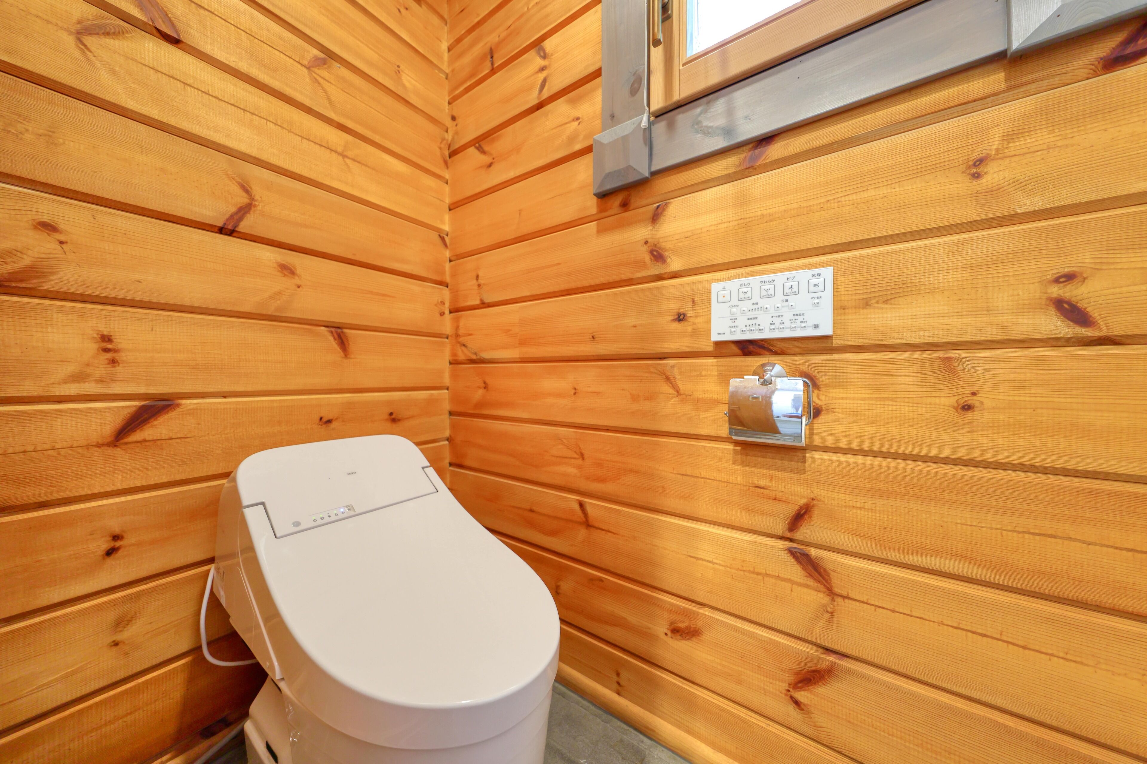 A wooden-paneled bathroom with a white toilet, a control panel, and a window.