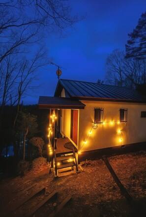 A house with a wooden staircase leading to a door, surrounded by a garden with a wooden fence and string lights.