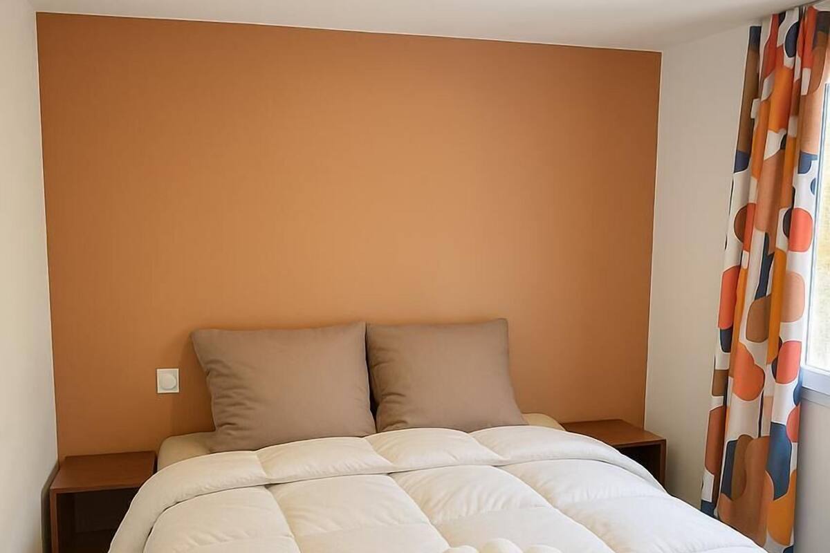 A neatly made bed with a beige duvet and pillows, a wooden nightstand, and a window with orange curtains.