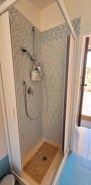Doccia con pareti piastrellate, soffione, doccetta e un piccolo ripiano con articoli da toilette.