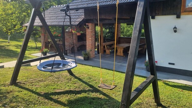 Un ensemble de jeux d’extérieur en bois comprenant une balançoire avec un siège en métal, un espace barbecue en briques et une terrasse en bois avec des sièges d’extérieur.