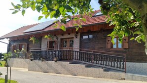 Une maison en bois avec des fondations en pierre, un balcon doté d’une balustrade en métal et un toit recouvert de tuiles rouges.