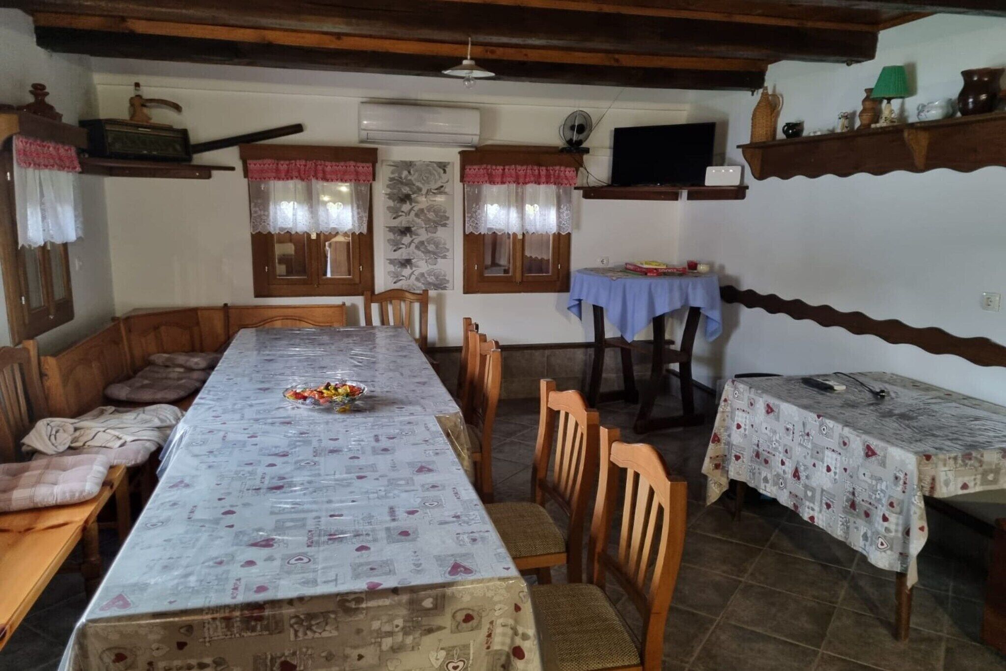 A dining area with a long table, wooden chairs, and a bench. There are two small tables with tablecloths, and a television on a shelf.