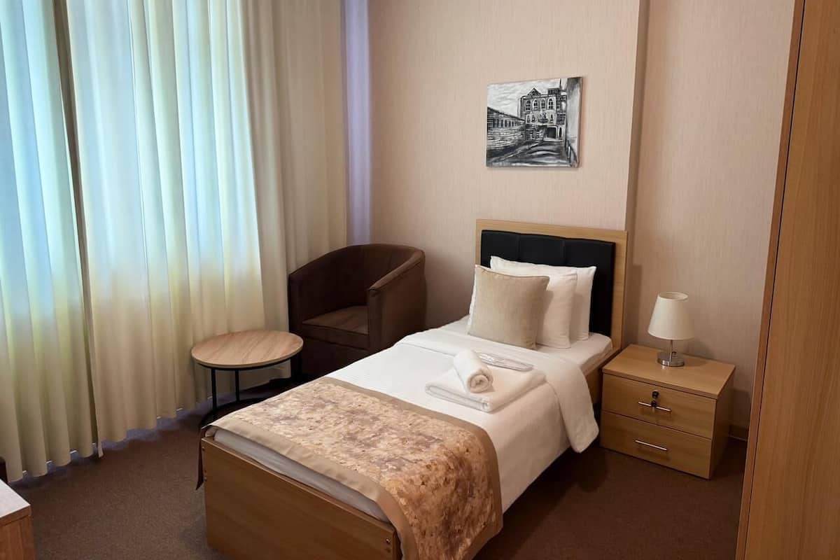 A hotel room with a bed, a chair, a small table, a nightstand, and a painting on the wall.