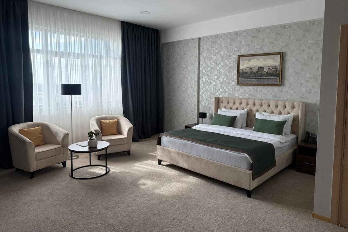 A hotel room with a large bed, two armchairs, a small round table, and a cityscape painting on the wall.