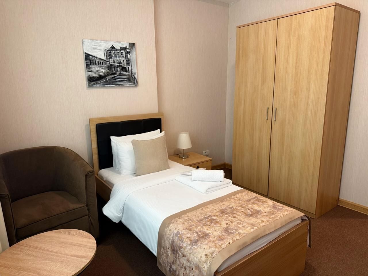 Habitación de hotel con cama, una silla, armario, mesita de noche con lámpara y un cuadro en la pared.
