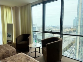 Quarto de hotel com uma grande janela, duas poltronas, uma mesa redonda pequena e um sofá.