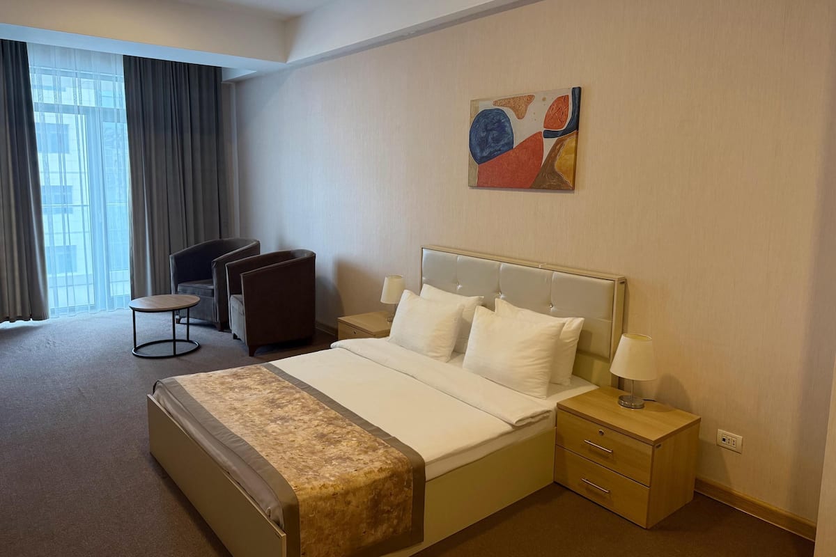 A hotel room with a bed, bedside tables, a chair, a small table, and a window with curtains.