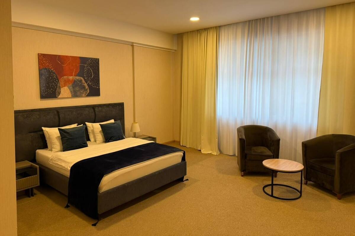 A hotel room with a bed, two armchairs, a small table, a nightstand, a lamp, and a painting on the wall.