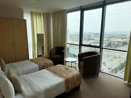 Quarto de hotel com duas camas, uma cadeira, uma mesinha e uma janela grande com vista para a cidade.
