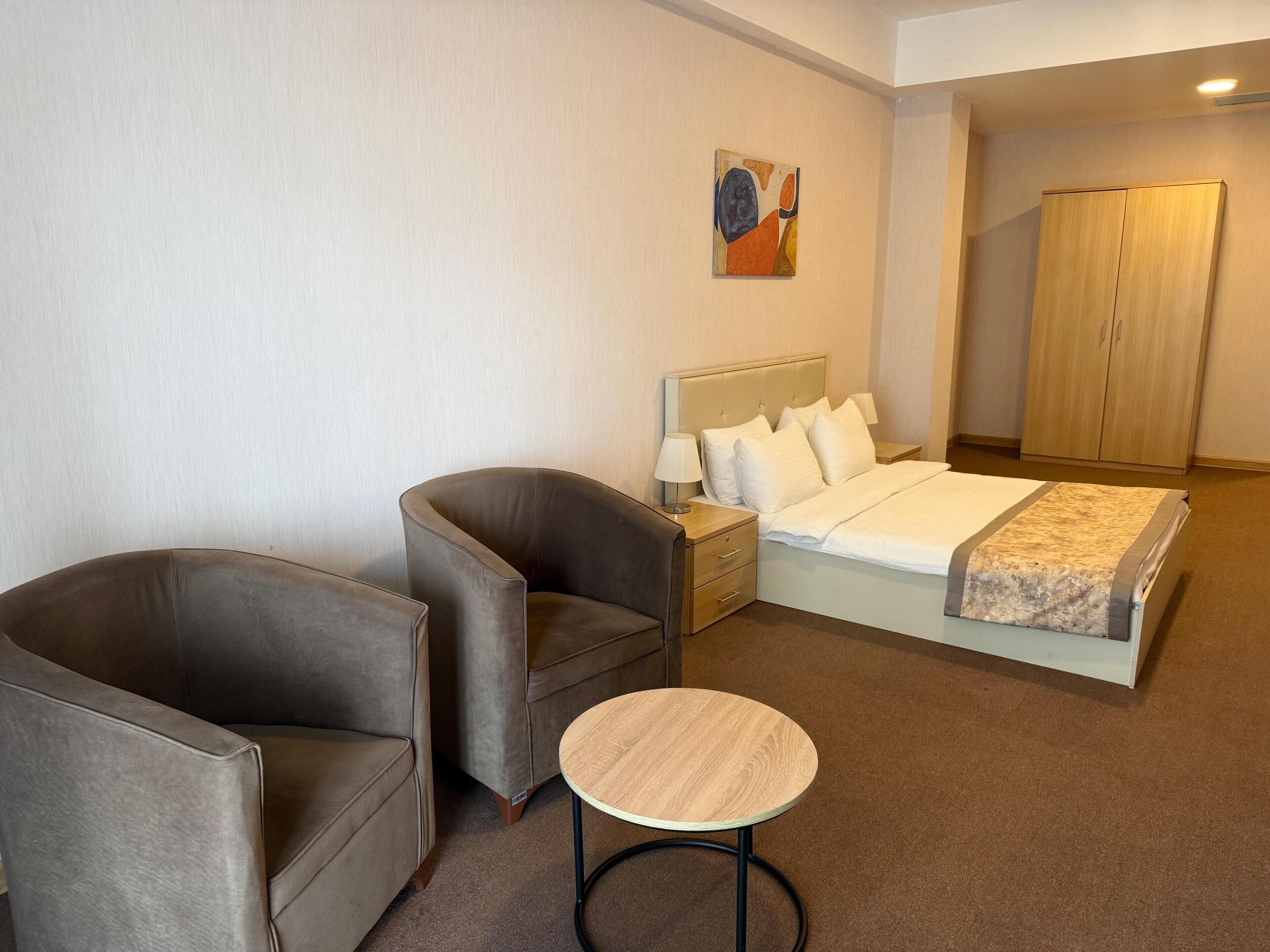 A hotel room with a bed, two armchairs, a small round table, a nightstand with a lamp, and a wardrobe.