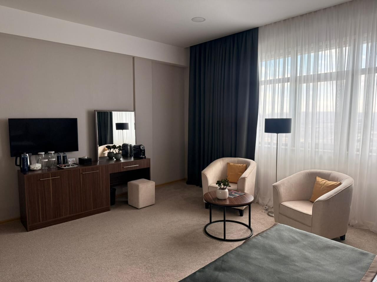 A modern hotel room with a flat-screen TV, a wooden desk, a sofa, a chair, and a large window with curtains.