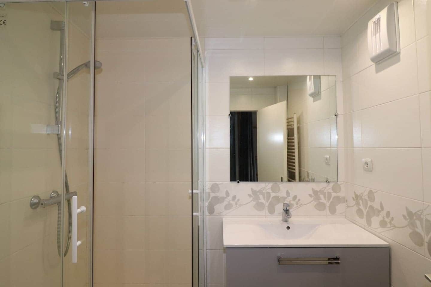 Une salle de bain avec une douche à parois de verre, un lavabo avec un miroir et un mur recouvert de carreaux décorés d’un motif floral.