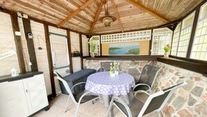 A covered patio with a stone wall, a table with chairs, and a view of the sea through a window.