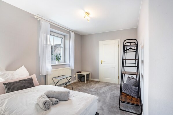 A bedroom with a bed, a window with curtains, a radiator, a small table, a ladder, and a door.