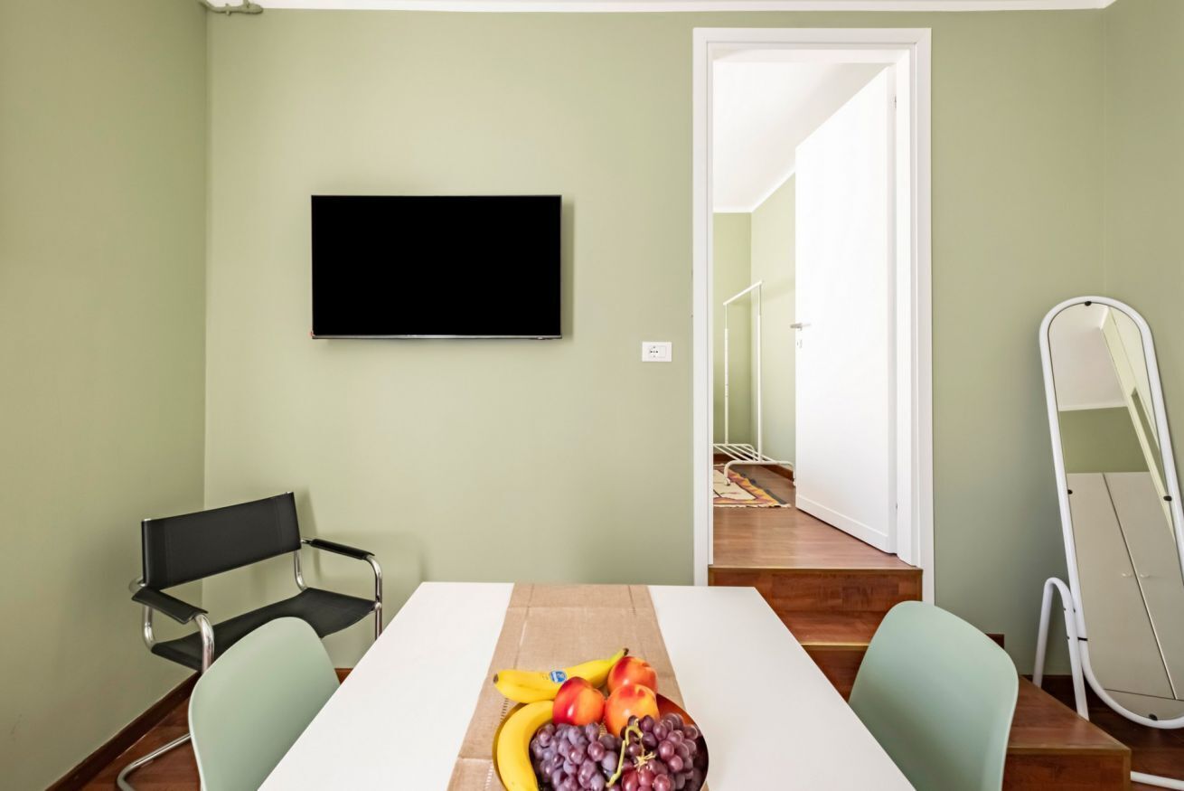 Una stanza con una TV a schermo piatto montata a parete, un tavolo con della frutta, due sedie e una porta che dà su un'altra stanza.