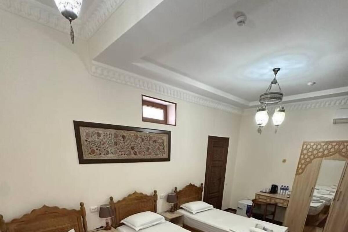 Habitación de hotel con tres camas individuales, cabecero de madera, espejo grande, escritorio con silla y alfombra roja.