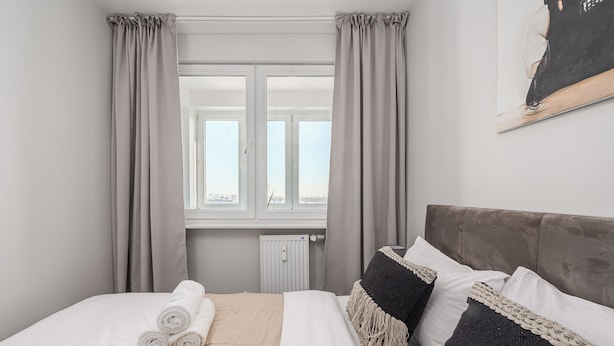 Ein ordentlich bezogenes Bett mit aufgerollten Handtüchern, einem Heizkörper und einem fenster mit Vorhängen.