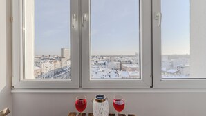 Ein Zimmer mit drei Fenstern, jedes mit Blick auf die Stadt. Ein Tisch mit zwei Gläsern mit roter Flüssigkeit und einem weißen Keramikbehälter.