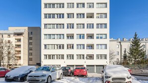 Ein hohes, mehrstöckiges Wohngebäude mit vielen Fenstern und Balkonen, umgeben von parkenden Autos und kahlen Bäumen.