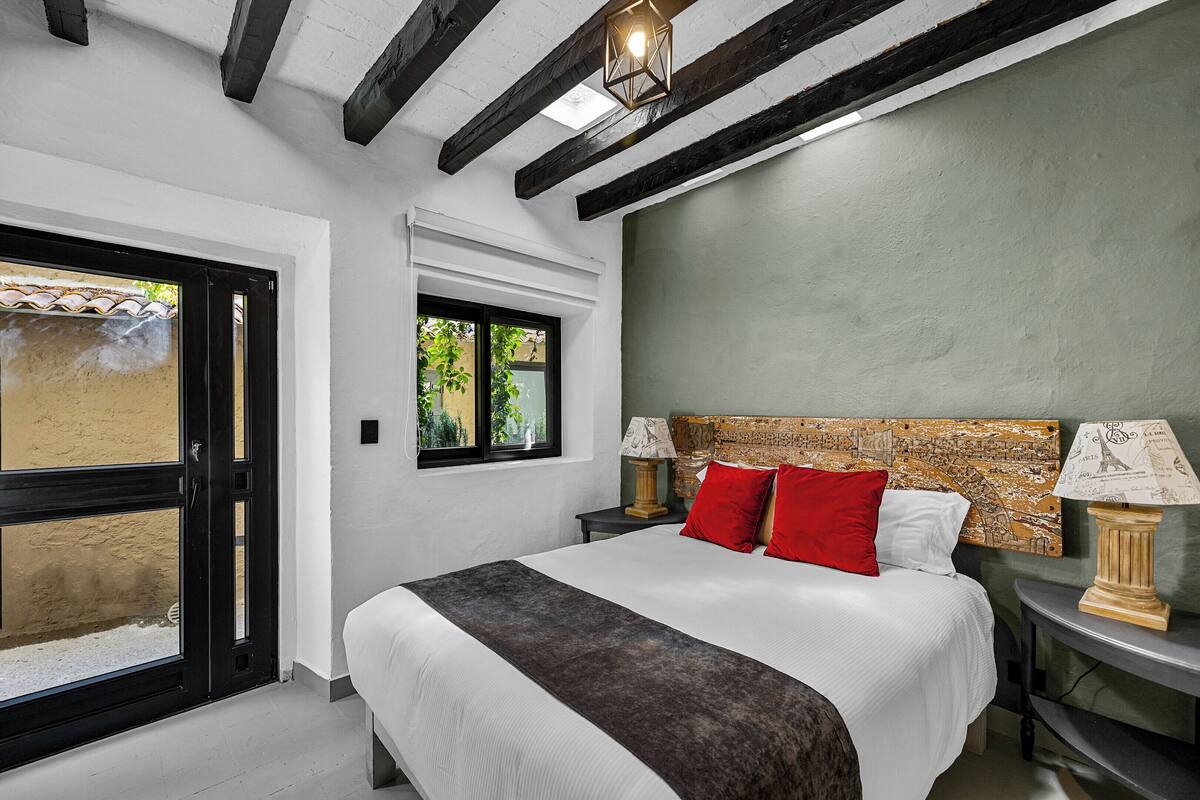 A bedroom with a bed, two bedside tables, a window, and a view of the outside.