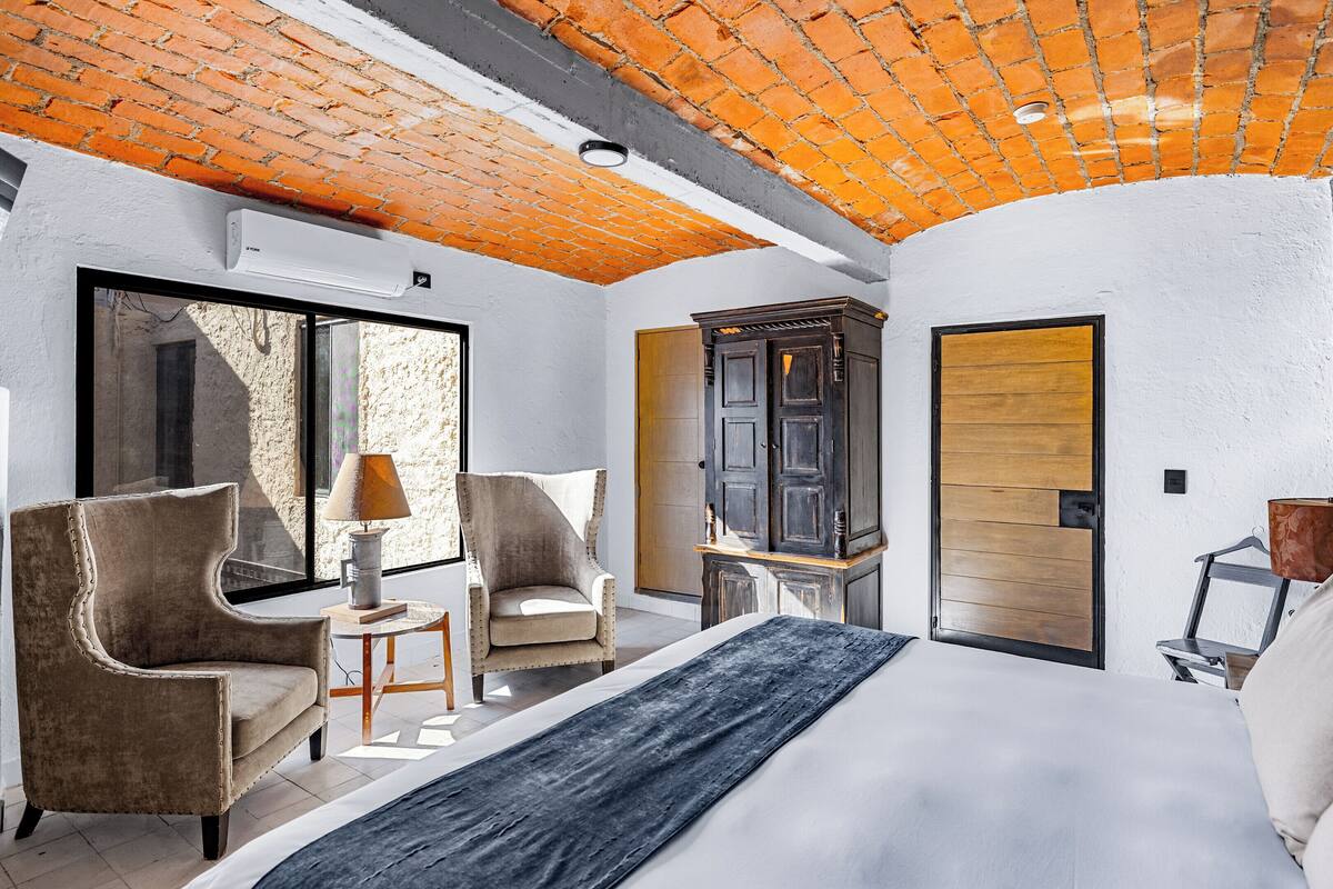 A bedroom with a large bed, two armchairs, a small table with a lamp, a wardrobe, and a sliding glass door to a balcony.