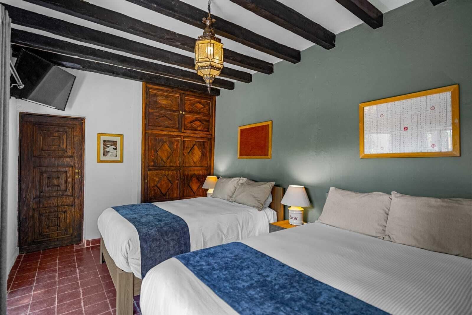 A room with two beds, a wooden door, a wall-mounted lamp, and a framed chart on the wall.