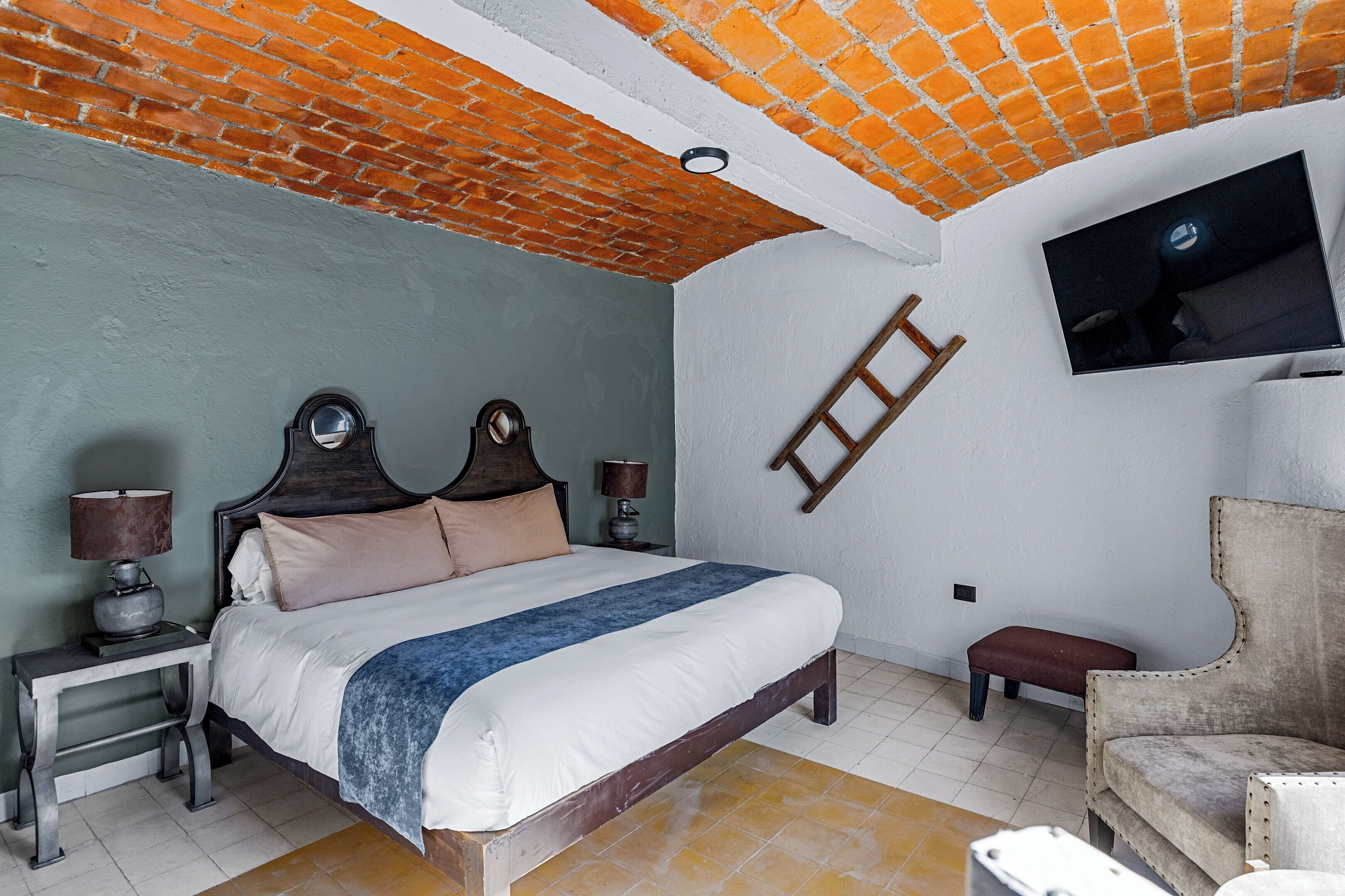 Quarto com teto de tijolos, cama com cabeceira de madeira, duas mesas de cabeceira com abajures, televisão montada na parede e uma cadeira.