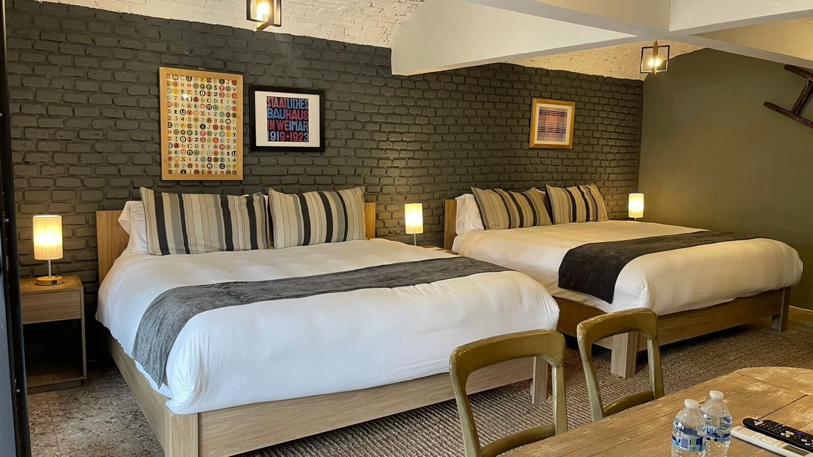 A hotel room with two beds, a small table, and a chair. There are wall-mounted lamps and a framed picture on the wall.