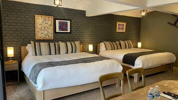 A hotel room with two beds, a small table, and a chair. There are wall-mounted lamps and a framed picture on the wall.