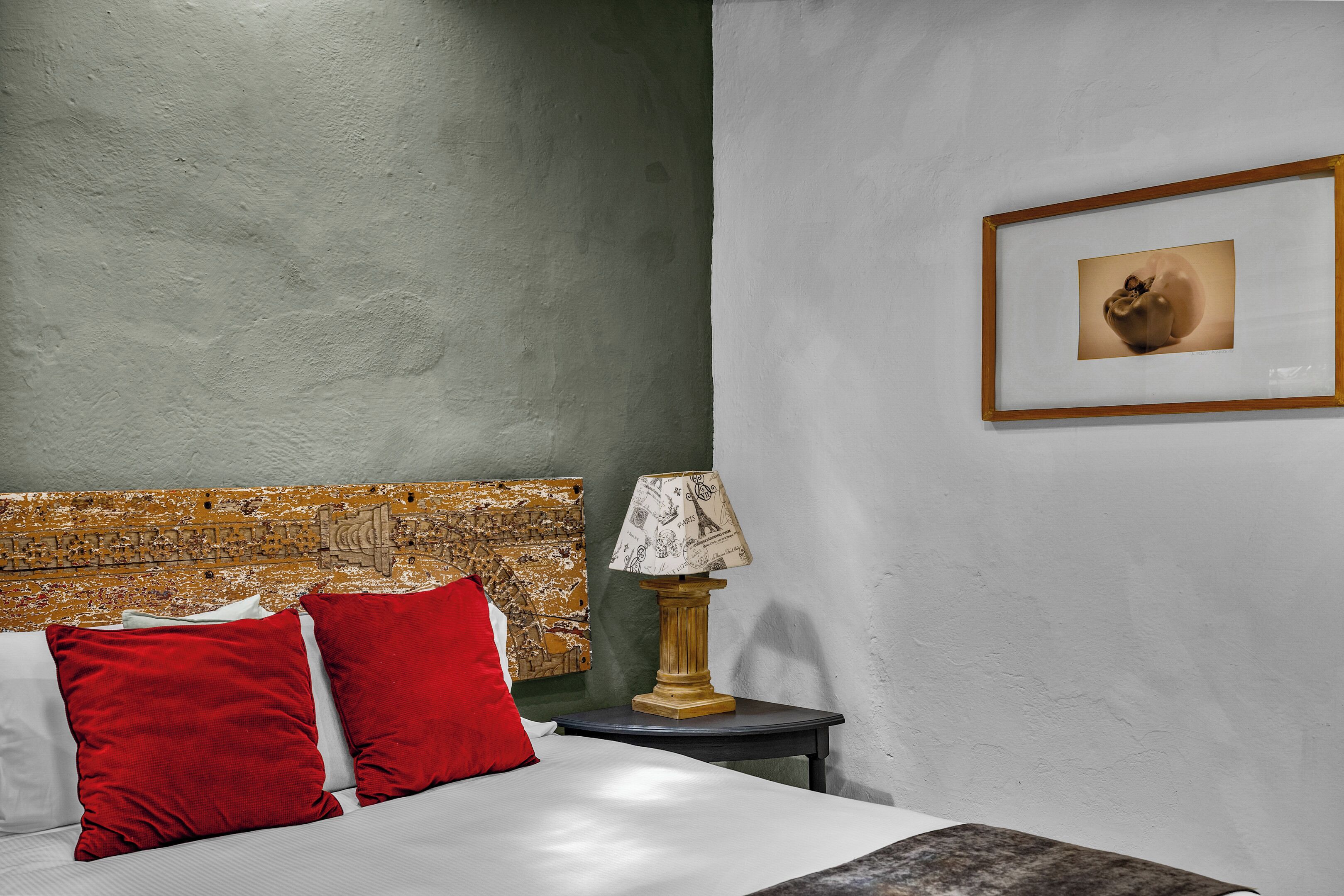 Quarto de hotel com uma cama, almofadas vermelhas, um abajur e um quadro na parede.