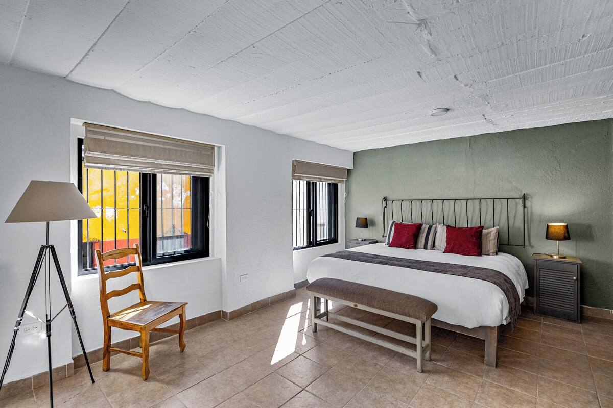 A bedroom with a bed, a chair, a bench, a lamp, and a window with blinds.