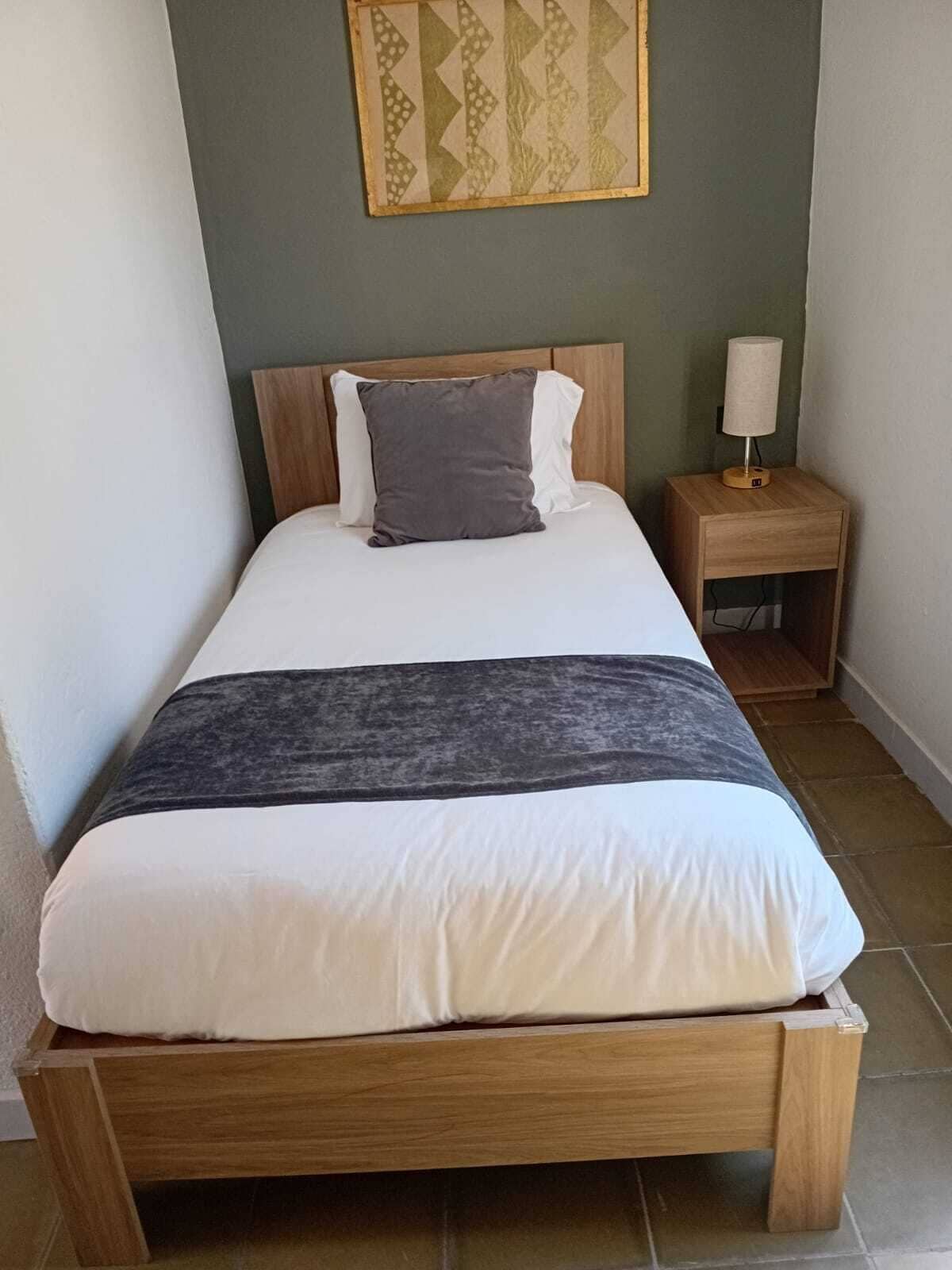 A single bed with a wooden frame, a bedside table with a lamp, and a patterned wall hanging.