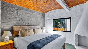 Un dormitorio con techo de ladrillo, paredes blancas, una cama con manta azul y una ventana con vista a la vegetación.