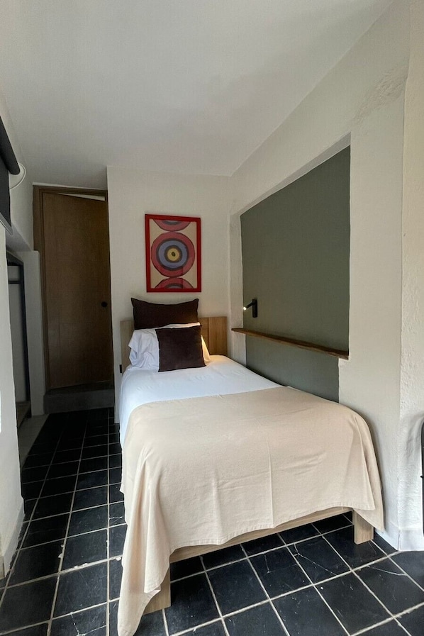 Une chambre d’hôtel étroite, avec un lit, une œuvre d’art accrochée au mur et une porte donnant sur une autre pièce.