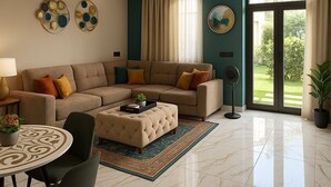 A modern living room with a beige sectional sofa, a round coffee table, and a tufted ottoman. There is a dining area with a table and chairs, a standing fan, and a large window with a view of a garden. - Swagatha The Beach House ECR (Kanchipuram District,ECR 603104)