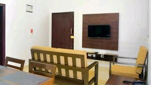 A room with a wooden sofa, a small table, a TV mounted on the wall, and a door. - VARSHA ENCLAVE TRINITY HOUSE (Mysore)