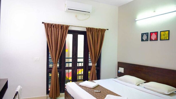 A hotel room with a bed, curtains, an air conditioner, and a wall with three framed pictures. - VARSHA ENCLAVE TRINITY HOUSE (Mysore)