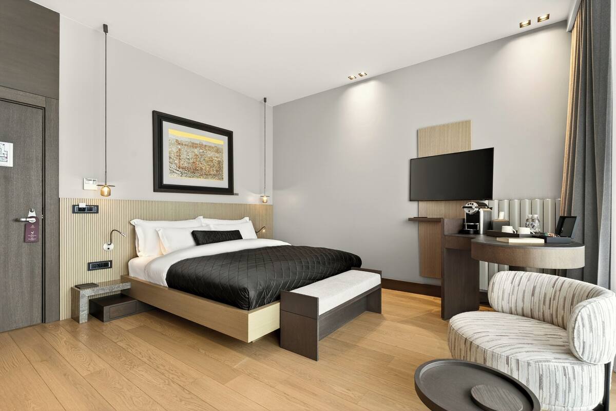 A modern hotel room with a bed, bedside table, chair, and a flat-screen TV.