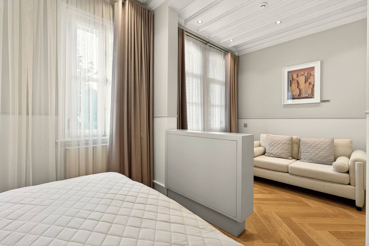 A modern hotel room with a large bed, a sofa, a radiator, and a cityscape painting on the wall.