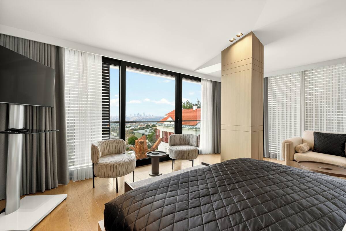 A modern hotel room with a large bed, two armchairs, a small table, and a sofa. The room has large windows with blinds, a flat-screen TV, and a view of a cityscape.