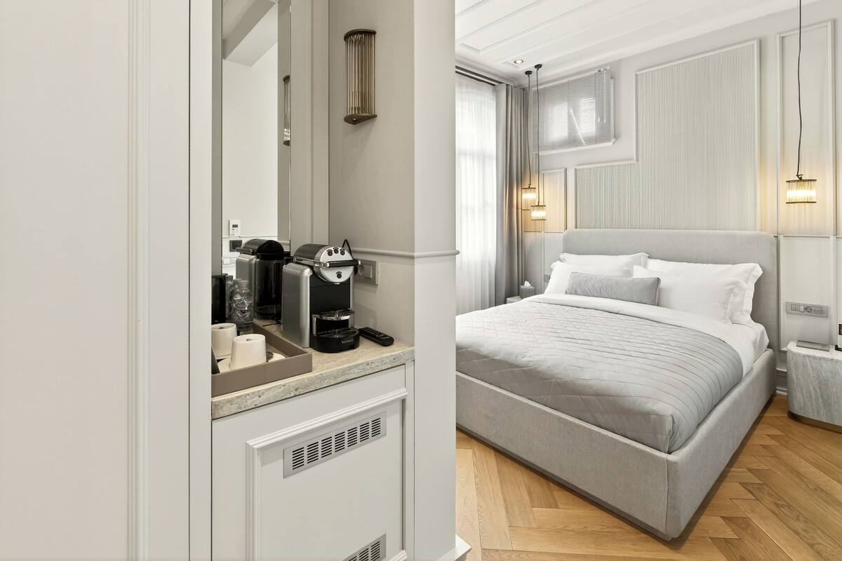 A modern hotel room with a large bed, a bedside table with a coffee maker, a mirror, and a view of the city through the window.