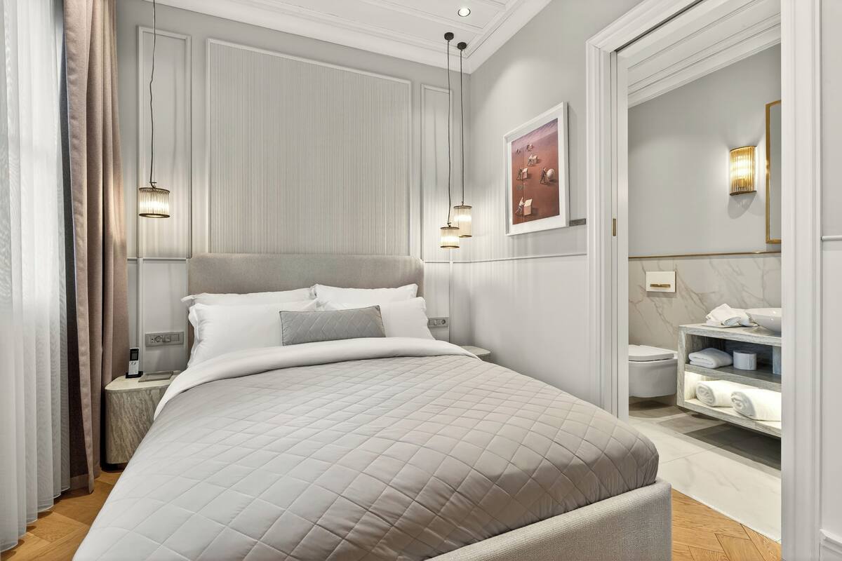 A modern hotel room with a large bed, bedside tables, a nightstand, a wall-mounted light fixture, a bathroom with a toilet, a sink, and a towel rack.