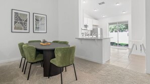 A modern dining area with a round table and green upholstered chairs, a kitchen with white cabinets, and a view of the outdoors through a glass door.