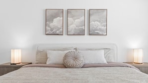 A bedroom with a large bed, two bedside lamps, and three framed cloud paintings on the wall.
