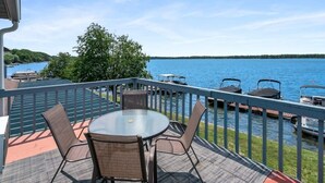 A deck with a round table and chairs, overlooking a lake with boats docked.