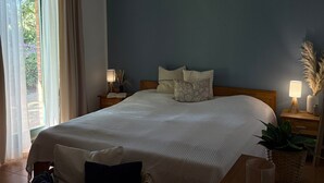 A bedroom with a large bed, a nightstand with a lamp, a window with curtains, and a view of greenery outside.