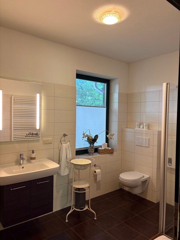 A modern bathroom with a white sink, dark cabinets, a small window with a view, a toilet, and a shower area.
