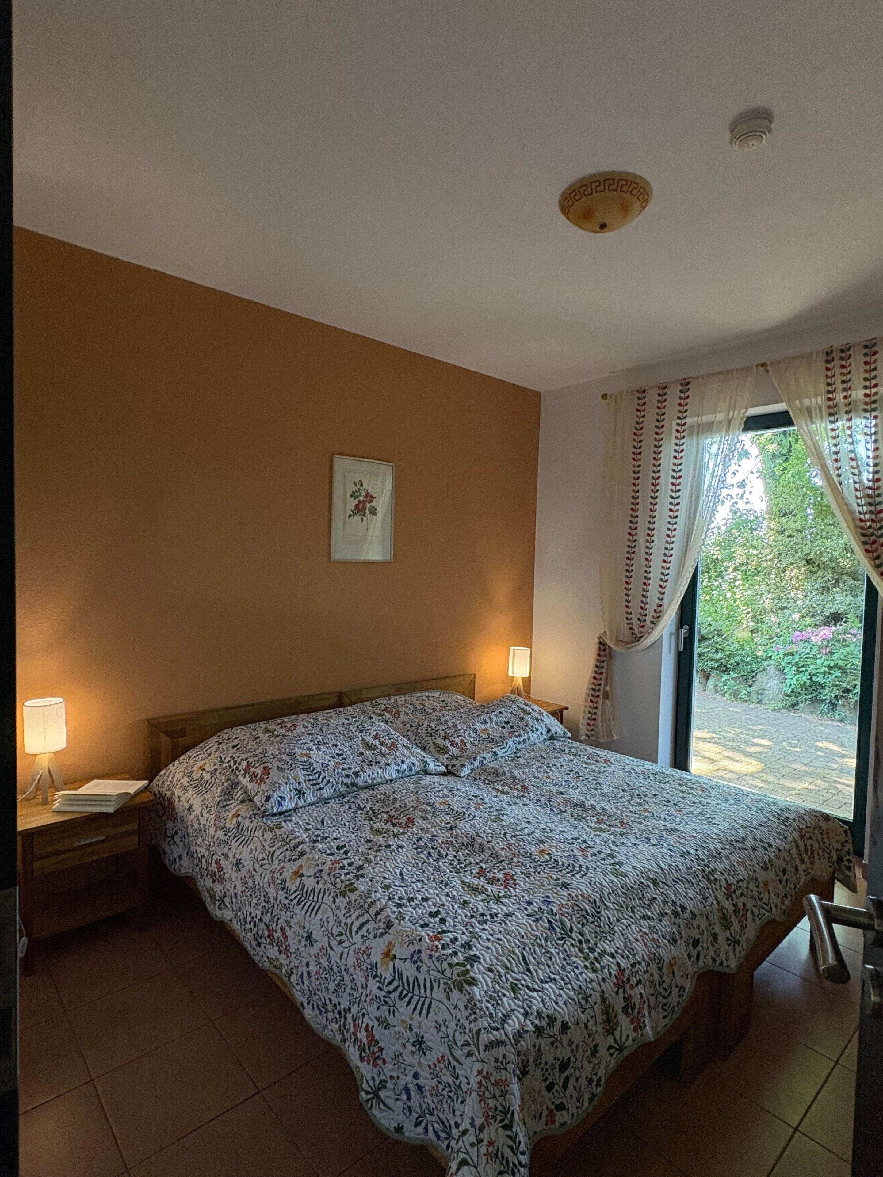 A bedroom with a large bed, two bedside lamps, a framed picture on the wall, and a window with curtains.