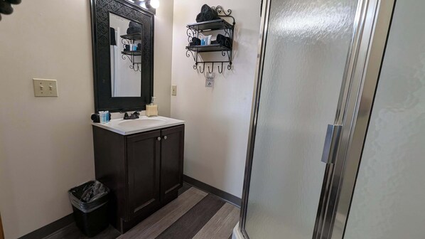 A bathroom with a mirror, sink, dark cabinets, a trash bin, a shower area, and a towel rack with toiletries.